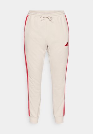Beige joggebukser med røde sidestriper, elastisk liv med snor, mansjetter på ankelen og en brodert logo på låret.