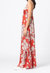 Červené maxi šaty s bílými květinovými vzory, s ramínky na tenké špagety, splývavý materiál a volánkový lem. Materiál vypadá lehký a hladký.