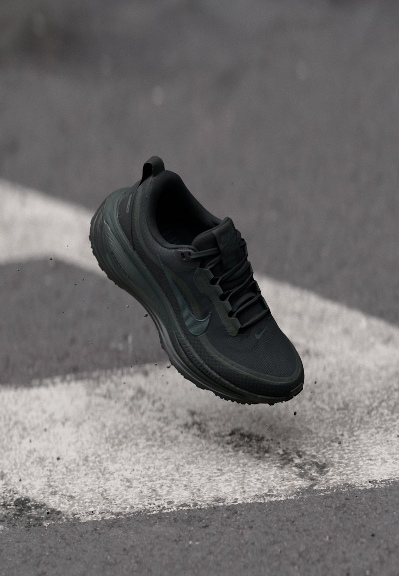 Scarpa da corsa nera Nike sospesa sopra l'asfalto con segni stradali bianchi e piccole particelle di terra attorno.