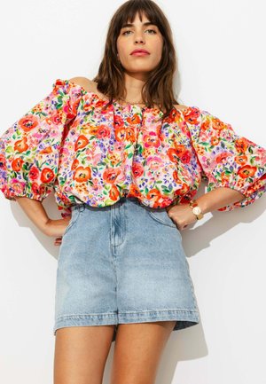 Femme portant un chemisier fleuri coloré à épaules dénudées avec des manches bouffantes et un short en jean bleu clair taille haute, debout les mains sur les hanches.