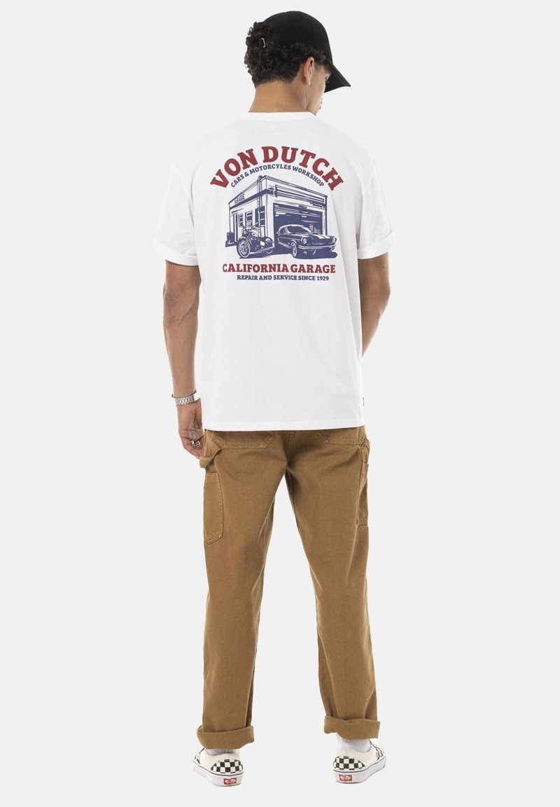 T-shirt blanche avec un graphique rouge et bleu au dos, présentant des voitures vintage et le texte "VON DUTCH CALIFORNIA GARAGE". Portée avec un pantalon cargo beige.