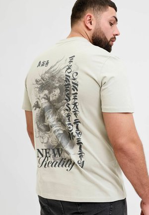 Homme aux cheveux foncés et barbe porte un T-shirt beige clair avec un grand motif de dragon noir et un texte stylisé « New Reality » dans le dos.