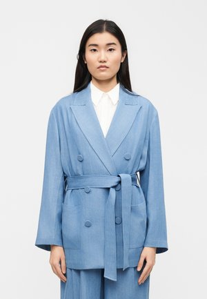 Jonge vrouw die een lichtblauwe, dubbelgeknoopte blazer met ceintuur draagt over een witte blouse met kraag, staand tegen een effen achtergrond.