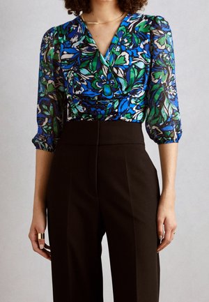 Kobieta ubrana w wysokie czarne spodnie i niebiesko-zielono-czarną kwiecistą bluzkę na zakładkę z trój-czwartego rękawa z bufkami.