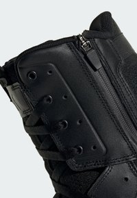 Primer plano de una zapatilla negra de caña alta con cuero texturizado, cremallera lateral y cordones negros pasados por ojales de metal.
