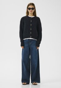 Tmavě námořnický pletený cardigan se zlatými knoflíky, nošený přes bílý top. V páru s širokými modrými džíny a černými žabkami.