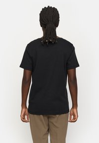 Black cotton t-shirt with a relaxed fit, short sleeves, and a crew neck, viewed from the back, paired with beige pants.