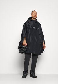 Črn vodoodporen pončo s kapuco, ki ima sprednji žep, v kombinaciji s temnimi hlačami in drži čelado v črni barvi. Gladek in lahek material.