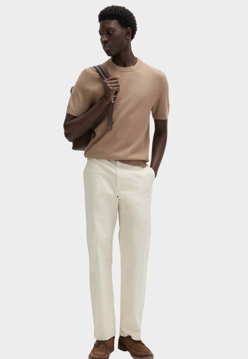 Ung mand iført en beige short-sleeve sweater, cremefarvede bukser, brune sko, der holder en brun rygsæksrem med den ene hånd og kigger til siden.