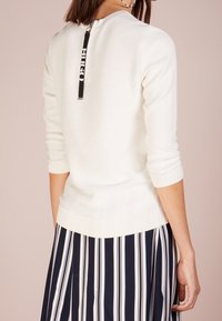 Donna che indossa un maglione bianco con texture e una cerniera nera con la scritta "HUGO" sulla schiena, abbinato a una gonna a righe verticali bianche e nere.