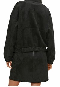 Vrouw in een zwarte lange mouwen jas met een elastische tailleband en een bijpassende zwarte rok met een zichtbare ritssluiting aan de achterkant.