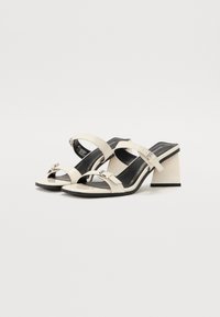 Sandalias de tacón abiertas en la parte delantera color beige con dos correas ajustables, con una plantilla negra y tacón cuadrado. Los acentos incluyen una pequeña hebilla decorativa.