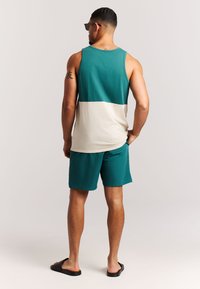 Camiseta de tirantes en verde azulado y beige, hecha de algodón, combinada con pantalones cortos en verde azulado. Modelo vistiendo sandalias negras, vista desde la parte trasera. Corte esbelto, detalles mínimos.