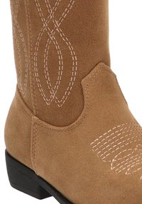 Bota de vaquero de ante marrón con patrones de costura intrincados y un tacón negro apilado. Presenta una punta redondeada y acentos decorativos en el cañón.