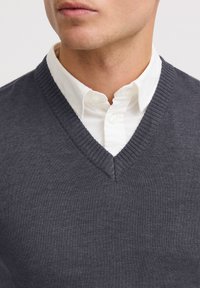 Pull en maille gris foncé à col en V sur une chemise à col blanc. Présente un col et un ourlet côtelés, une texture minimale et une coupe près du corps.
