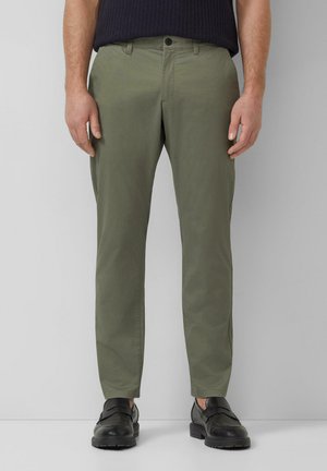 Man draagt olijfgroene broek met rechte pijpen, zwarte loafers en een donkerblauwe trui, staand tegen een effen grijze achtergrond.