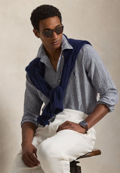 Άνδρας που φοράει ριγέ πουκάμισο με κουμπιά, με μπλε πουλόβερ ριγμένο στους ώμους, λευκό παντελόνι, ρολόι και γυαλιά ηλίου, καθισμένος σε ξύλινο σκαμπό.