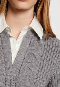Pull en tricot gris avec un col en V, mettant en avant des motifs en côtes et en torsades, superposé à une chemise blanche à col.