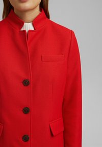 Rode jas met gestructureerde stof, hoge kraag, vijf donkere knopen en een borstzak. Voorzien van zijzakken en gedetailleerde stiksels.