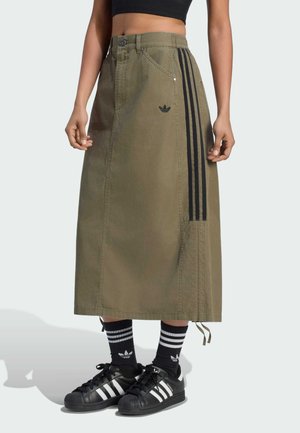 Donna che indossa una gonna midi verde oliva con strisce nere, scarpe da ginnastica nere Adidas e calzini neri con strisce bianche, in piedi su sfondo chiaro.