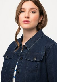 Giacca in denim blu scuro con colletto classico, due tasche sul petto e bottoni in metallo. Texture liscia con leggere sfumature di scoloritura.