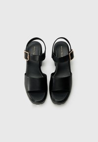 Schwarze Ledersandalen mit offenem Zeh und verstellbarem Knöchelriemen. Sie verfügen über einen klobigen Absatz und eine glatte Textur, die durch eine goldene Schnalle akzentuiert wird.