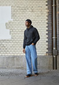 Svart hettegenser med frontlomme og snorer, sammen med lyseblå vide jeans. Ensfarget med minimal merkevare, står mot en murvegg.