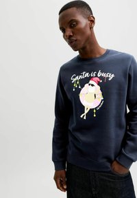 Muž v námořnickém svetru se Santou, který se povaluje na donutu, nosí sluneční brýle a má nápis "Santa má plné ruce práce" a vánoční světýlka.