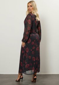 Vestido negro transparente con patrón floral rojo, mangas largas y un diseño fluido. Presenta un corte a media longitud y espalda descubierta. Tacones altos negros.