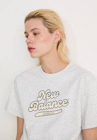 T-shirt en coton gris avec un logo "New Balance Athletics" en crème et en marine. Manches courtes et col rond.
