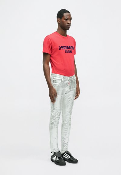 Punane t-särk, millel on tekst "DSQUARED2 MILANO", kombineeritud läikivate hõbedaste kitsaste teksade ja mustade sportlike kingadega, millel on valged triibud.