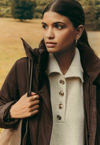 Pruun puffer-jakk kõrge kaelusega ja läikivate nuppudega, kihiline hele tekstuuriga sviiter. Kuldne sõrmus ja kõrvarõngad täiustavad riietust.