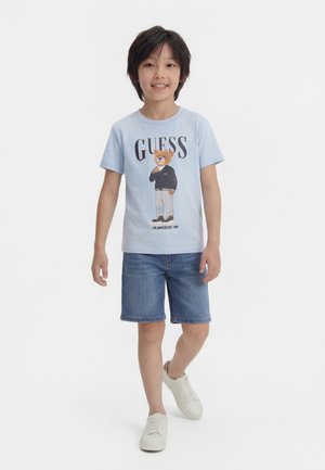 Ung dreng smilende, iført lyseblå T-shirt med bjørneprint og teksten "GUESS", denimshorts og hvide sneakers, stående mod en ensfarvet hvid baggrund.