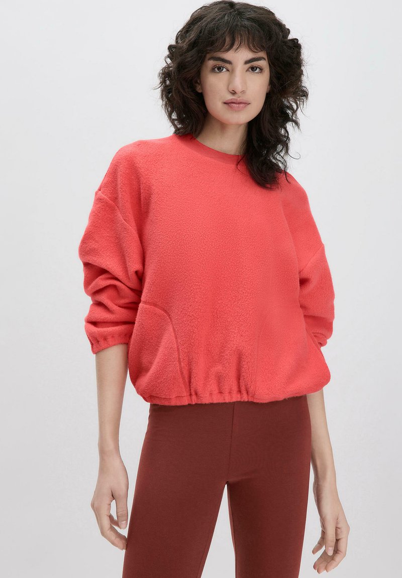 Coral Fleece Sweatshirt mit Rundhalsausschnitt, überschnittenen Schultern und elastischem Saum. Kombiniert mit dunkelroten Leggings. Weiche Textur.