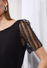 Top negru cu mâneci din material transparent, volanate, cu buline albe. Țesătura este fină și moale, creând o textură stratificată.