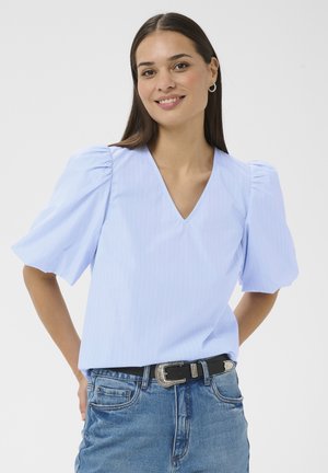Jeune femme souriante, portant un chemisier bleu clair à col en V avec des manches courtes bouffantes et un jean bleu avec une ceinture noire.