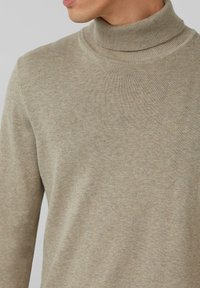 Beżowy golf wykonany z miękkiej tkaniny, z prążkowanym kołnierzem i gładką fakturą, charakteryzujący się minimalnym przeszyciem i dopasowanym krojem.