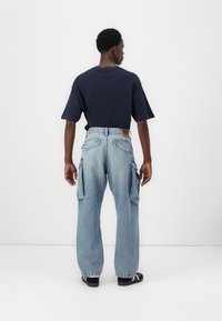 Μπλε τζιν cargo παντελόνι με υπερμεγέθη εφαρμογή, διαθέτοντας μεγάλες πλαϊνές τσέπες και μεσαία μέση. Συνδυασμένο με ένα σκούρο μπλε πουκάμισο κοντό μανίκι.