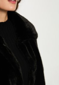 Cappotto nero in pelliccia sintetica con un ampio colletto e una texture morbida, abbinato a un maglione nero a costine sotto. Focus sulla lucentezza e sul calore lussuosi.