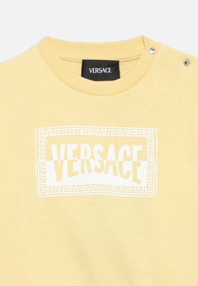 Жълта блуза с кръгло деколте с бяло лого на Versace в правоъгълна рамка с гръцки ключ и два сребърни копчета на рамото.