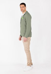 Hombre de pie de lado, vistiendo una chaqueta verde, pantalones beis, zapatillas blancas y una camiseta blanca, frente a un fondo blanco liso.