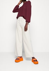 Haut à manches longues bordeaux avec poignets froncés, associé à un jean crème à jambes larges, et des sandales à plateforme orange, dotées de deux brides et d'une semelle texturée.