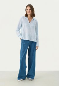 Camicia in lino azzurro chiaro con scollo a V e maniche lunghe, abbinata a jeans in denim blu a gamba larga e scarpe argentate, su uno sfondo neutro.