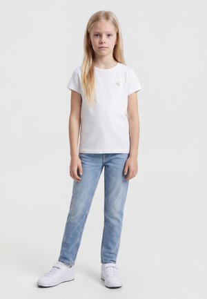 Chica rubia de pie, vestida con una camisa blanca de manga corta, vaqueros azul claro y zapatillas blancas contra un fondo claro y liso.