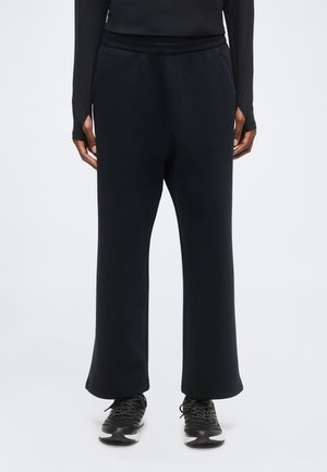 Persona que lleva pantalones negros sueltos, camiseta negra de manga larga y zapatillas negras con suelas blancas sobre un fondo blanco.