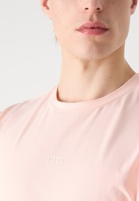 Lys rosa crew-neck t-skjorte laget av mykt stoff, med en subtil "BOSS"-logo i hvitt på brystet. Glatt tekstur, minimalistisk design.