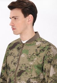 Giacca bomber in camouflage con un mix di motivi verdi e marroni, chiusura con zip e collo a costine. Tessuto dalla texture liscia.