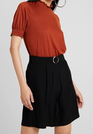 Vrouw draagt een roestrood kortemouwtop met gerimpelde kraag en zwarte, hooggetailleerde shorts met riem en ronde gesp.
