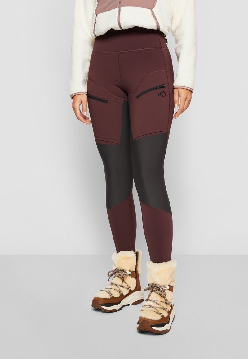 Frau in zwei-tonigen burgunder- und schwarzen Leggings mit Reißverschlusstaschen, cremefarbener Fleecejacke und beigen, fellgefütterten Winterstiefeln.