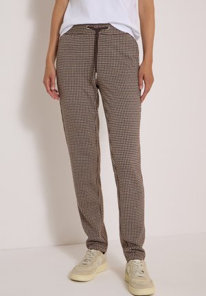 Personne portant un pantalon marron et beige à motif pied-de-poule, une chemise blanche et des baskets beiges, debout contre un mur clair et uni.